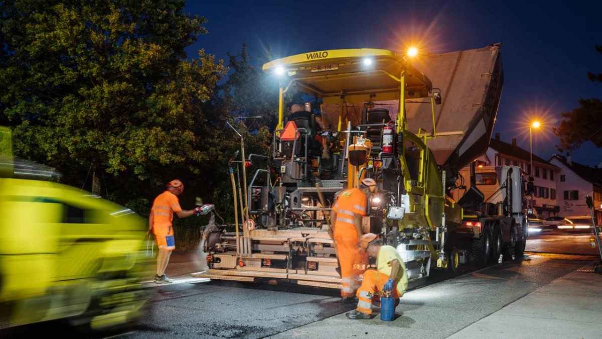 Grosse Maschine auf einer Strasse bei Nacht und Bauarbeiter, die die Strasse teeren