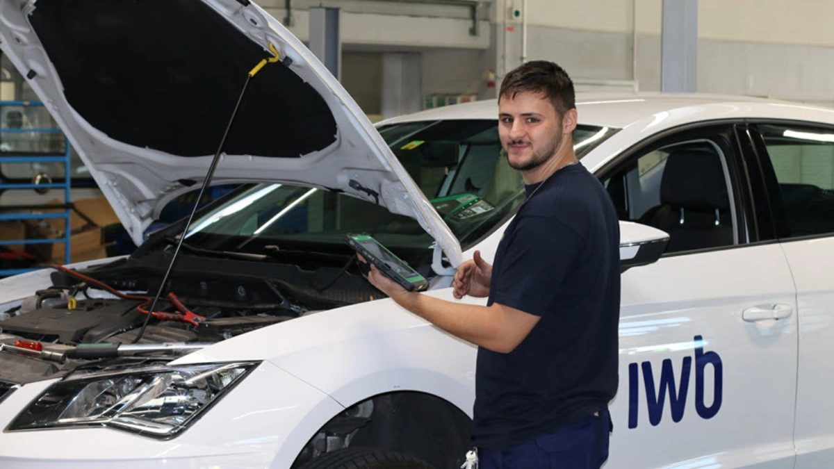 Ein junger Mann steht in einer Auto-Werkstatt. Vor ihm ist ein weisses Auto mit geöffneter Motorhaube.