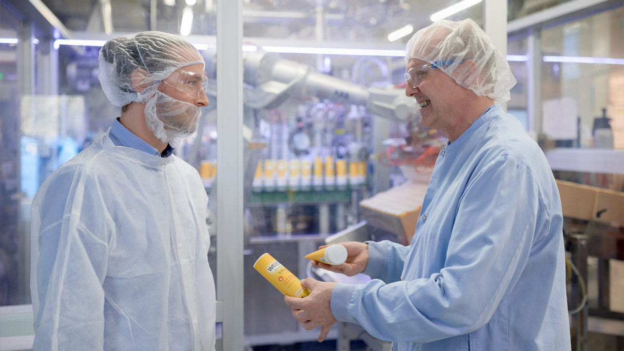 Zwei Männer in Schutzkleidung unterhalten sich in einer Produktionsumgebung, einer hält eine Sonnencreme in der Hand.