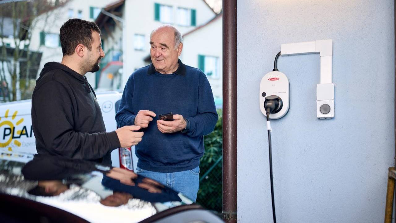 Zwei Personen stehen neben einer Wallbox an einem Einfamilienhaus und sprechen über das Laden eines Elektroautos mit Solarstrom.