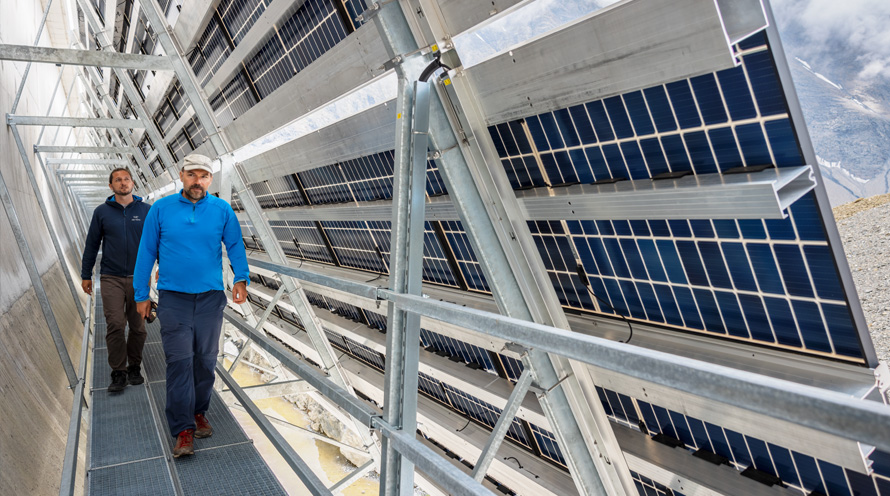 Zwei Männer laufen auf einem Metallsteg hinter einer Solaranlage hindurch.