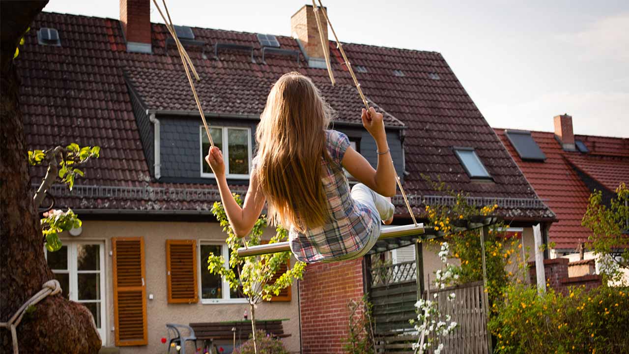 Ein Mädchen sitzt auf einer Schaukel, im Hintergrund ist ein Reihenhaus zu sehen